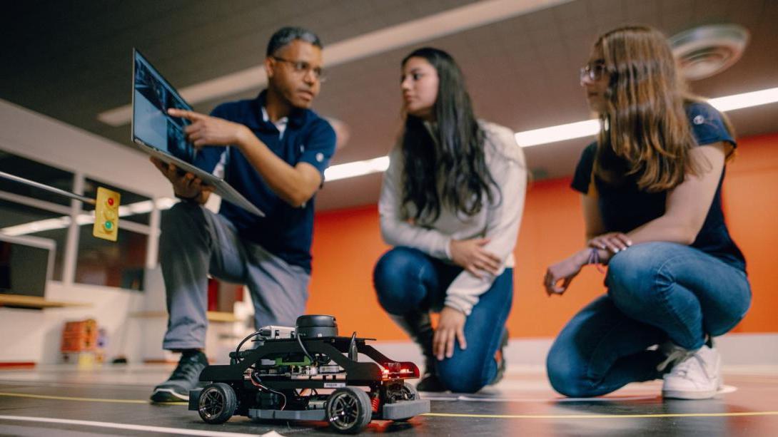 A Kettering professor points to a laptop as two students look on. In front of them is an autonomous electric vehicle